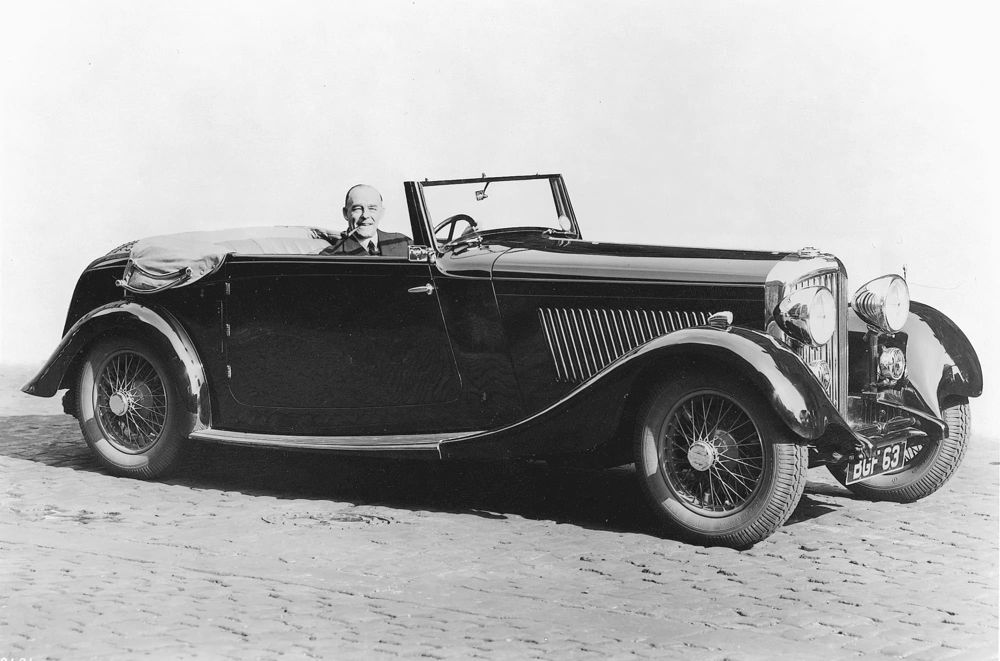 A black and white archive photograph of Alexander Duckham at the wheel of his first Bentley, a 1934 3½-litre convertible, on a cobbled street