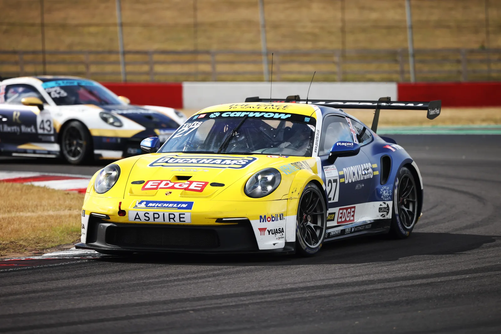 Jacob Tofts's number 27 Duckhams Porsche 911 GT3 Cup, yellow and blue, leading through a corner at Donington Park.
