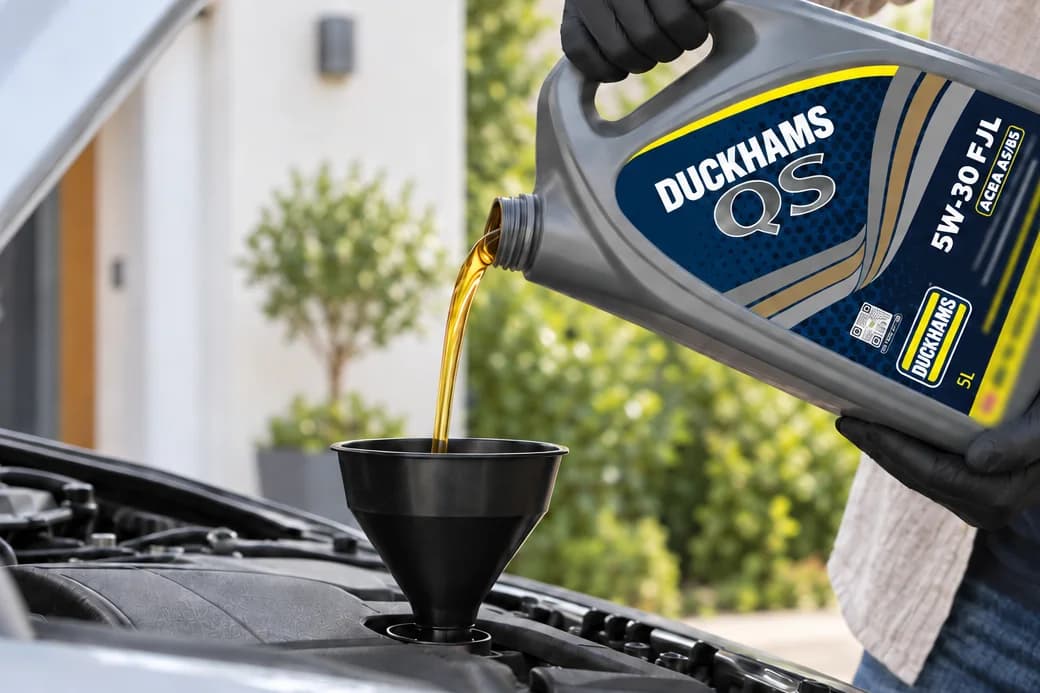A driver in black gloves pouring Duckhams QS 5W-30 into an engine through a funnel, on a driveway.