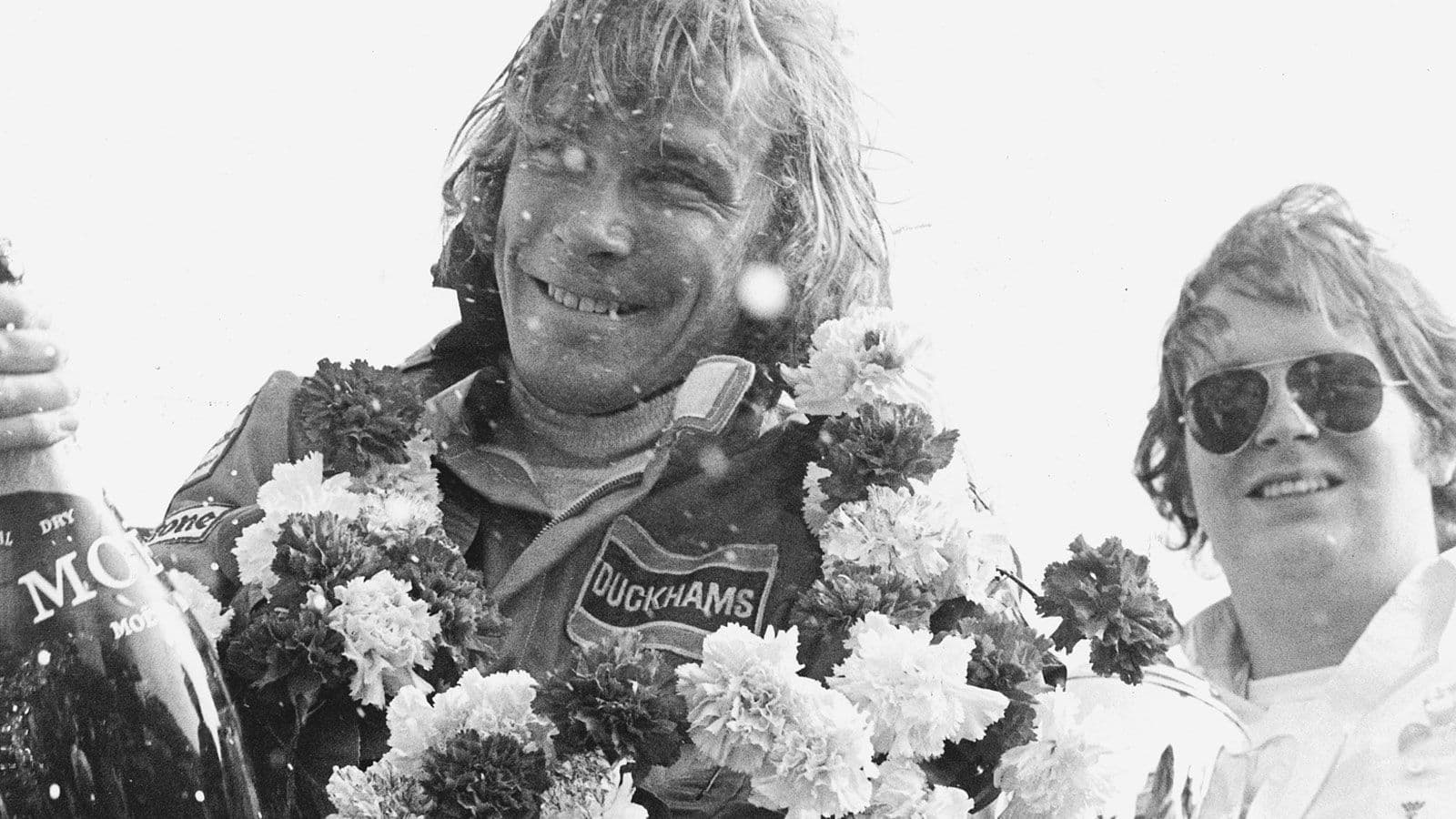 A garlanded racing driver in Duckhams overalls smiling beside a woman in sunglasses.